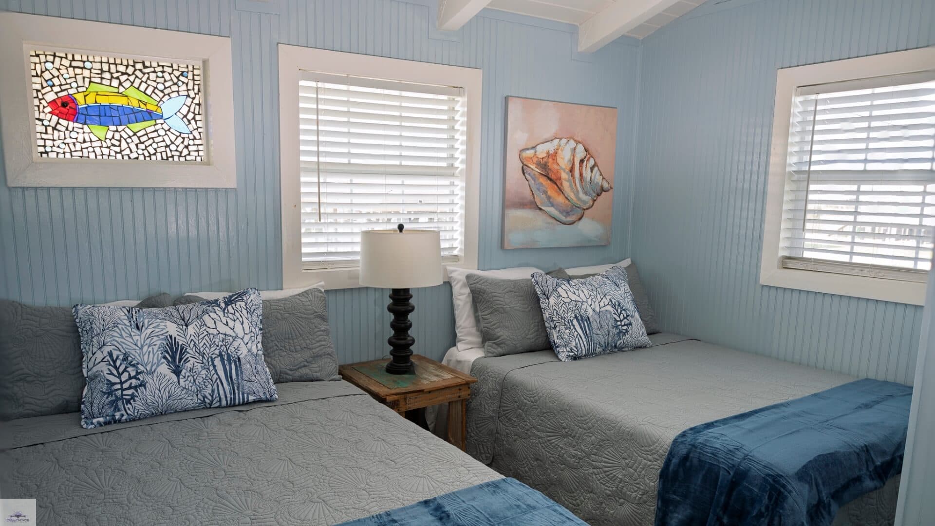 A cozy bedroom featuring two beds, a lamp, fish-themed stained glass, and sea shell artwork on light blue walls.