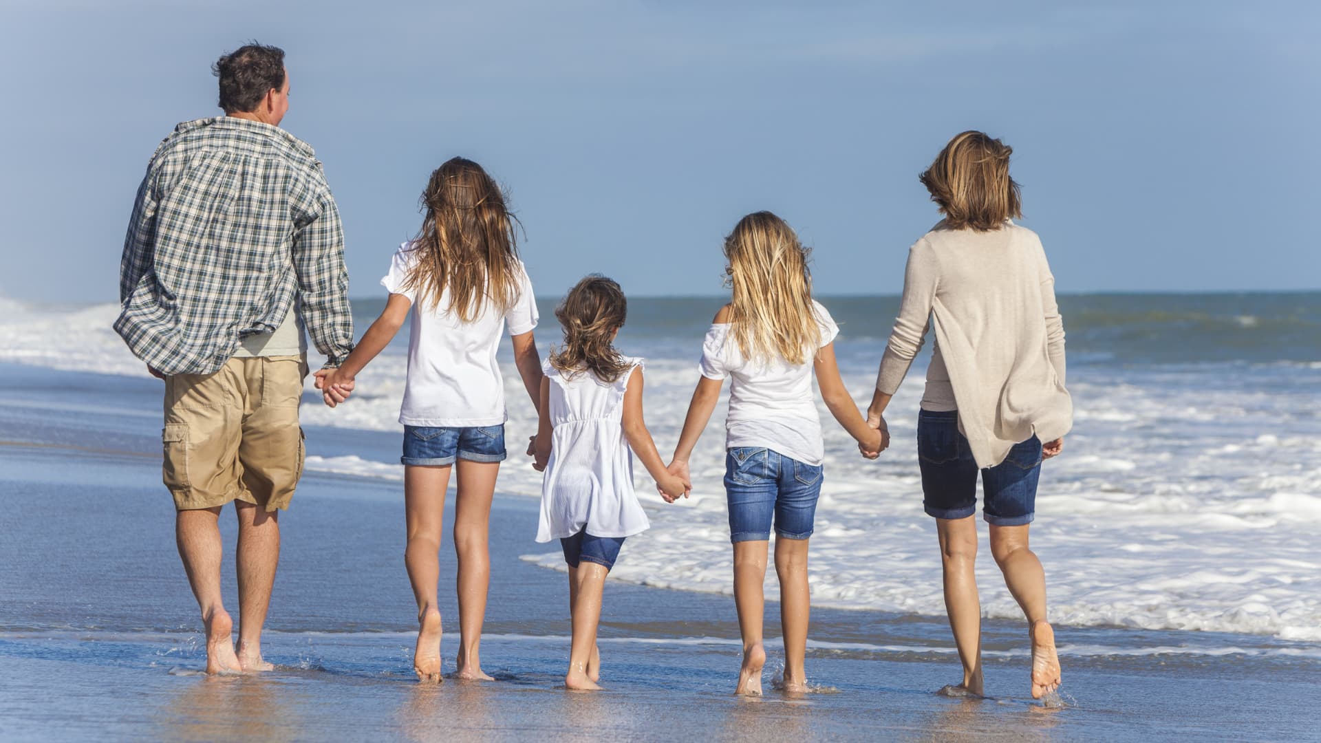 A family of five walks hand-in-hand along a sandy beach by the ocean.