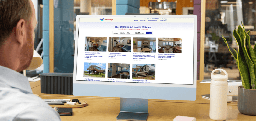 A person seated at a desk views the Blue Dolphin Inn website showcasing rooms and rates on a computer monitor.