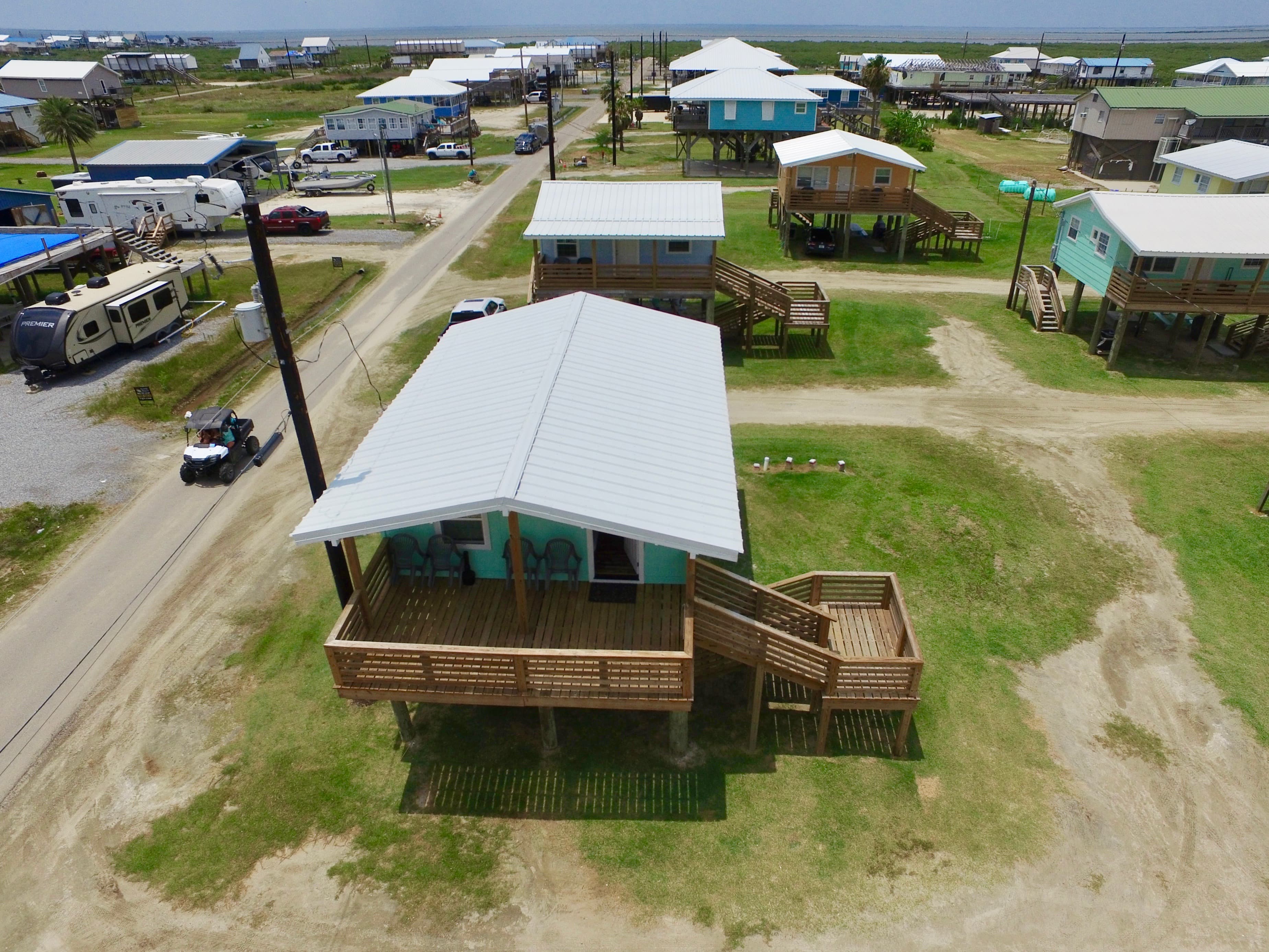 An aerial view of a stilt house with a white roof and large wooden deck, surrounded by other coastal homes and a street.