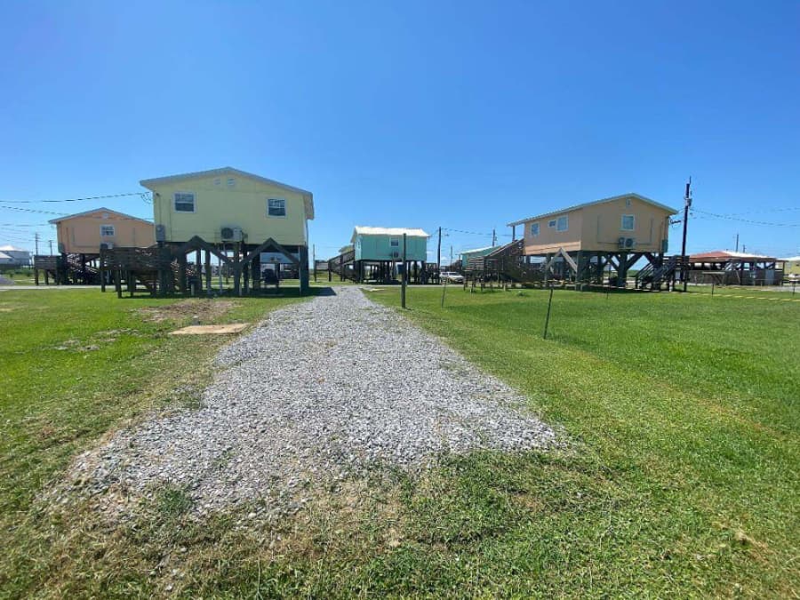 Two pastel-colored houses on stilts with a gravel pathway in between, set against a clear blue sky.