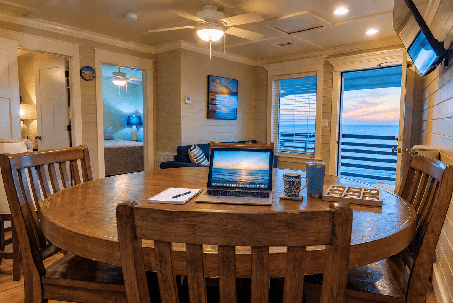 A cozy dining area features a round wooden table, a laptop, and a view of the ocean at sunset through large windows.