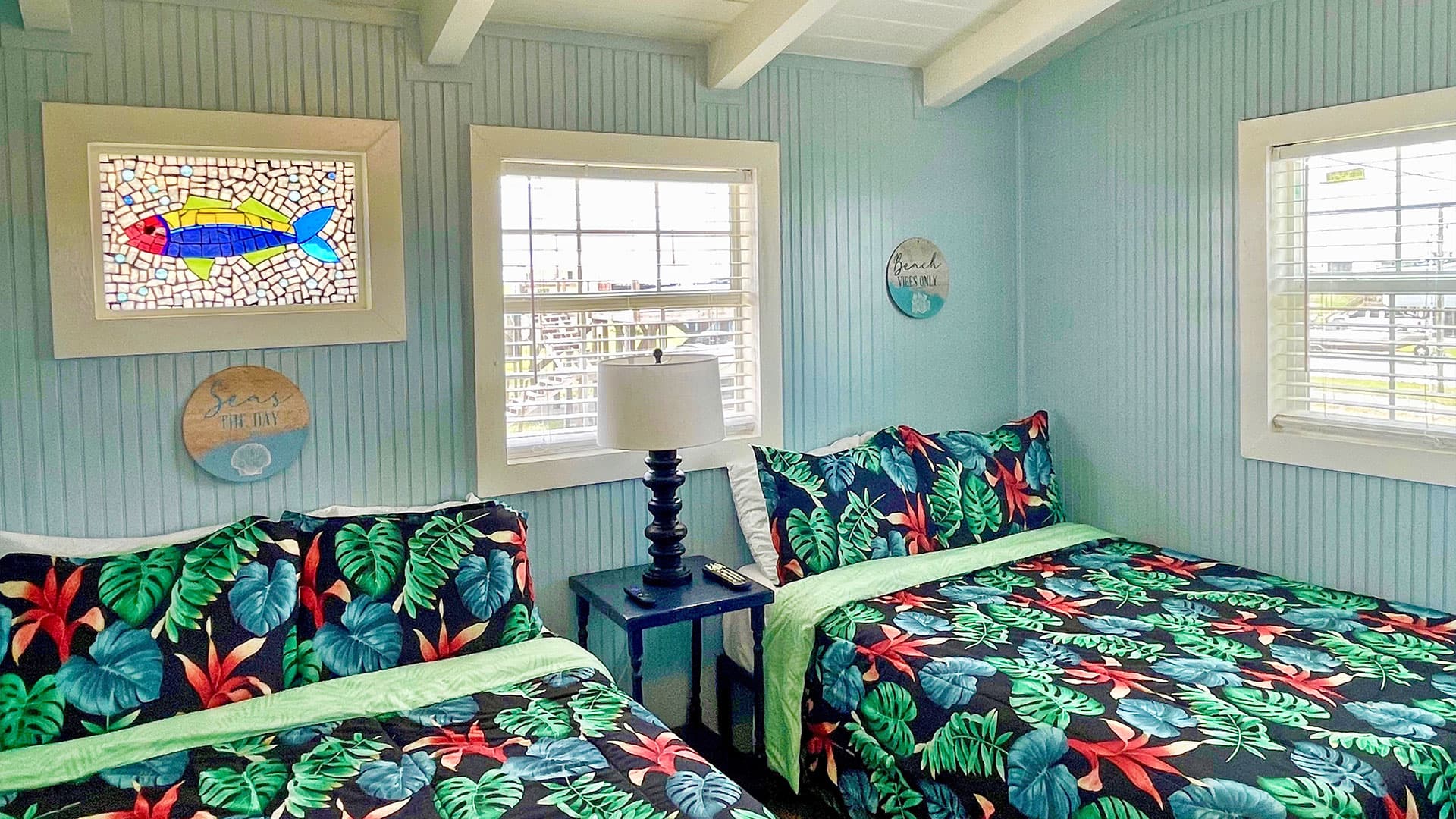 A bright, coastal-themed bedroom features two beds with tropical-patterned linens, a lamp on a small table, and colorful windows with decorative glass.