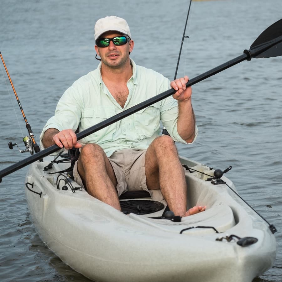 A man in a kayak holds a paddle and fishing rod while sitting in calm waters.