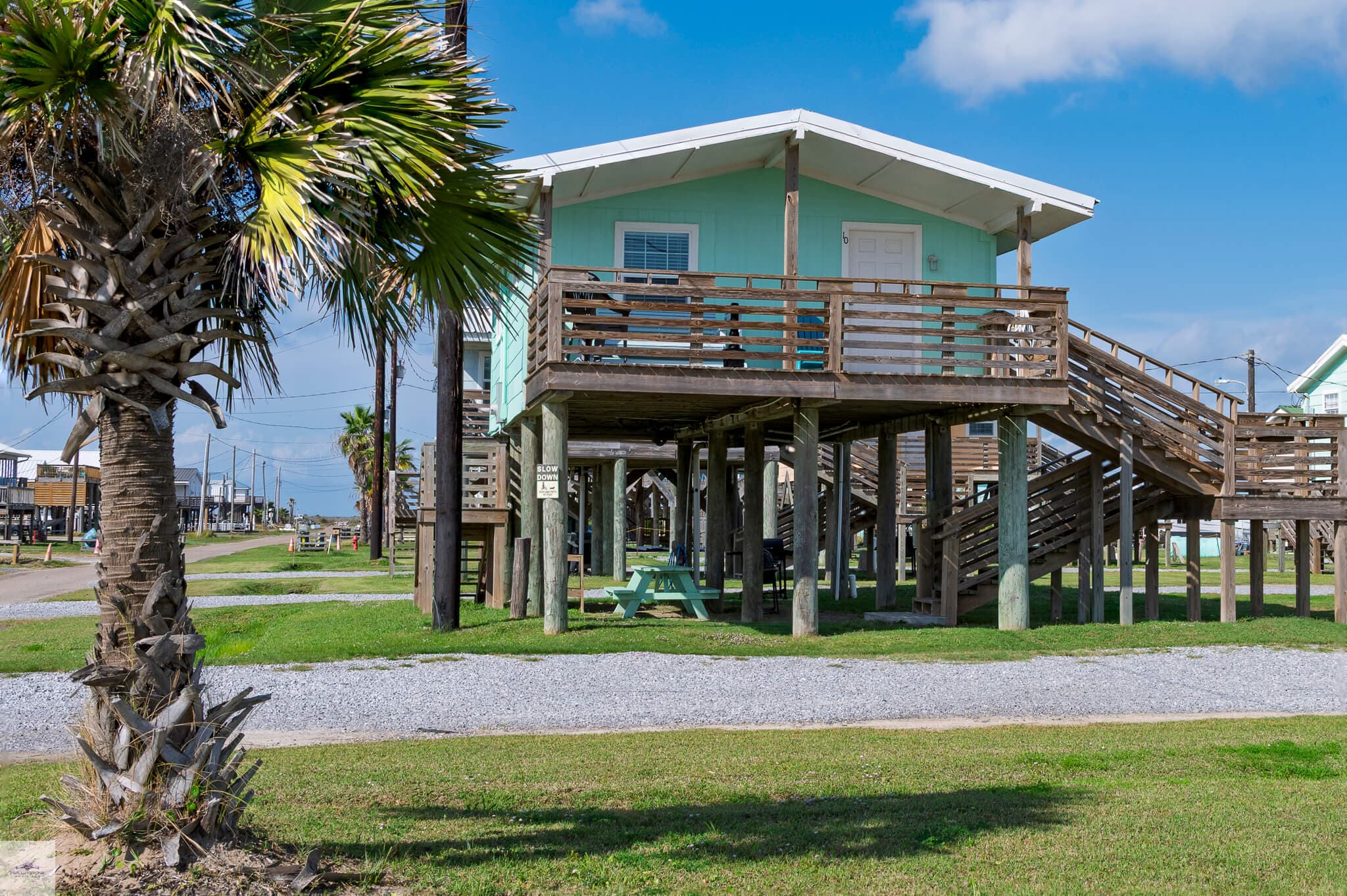 A teal beach house elevated on stilts surrounded by palm trees and a clear blue sky.