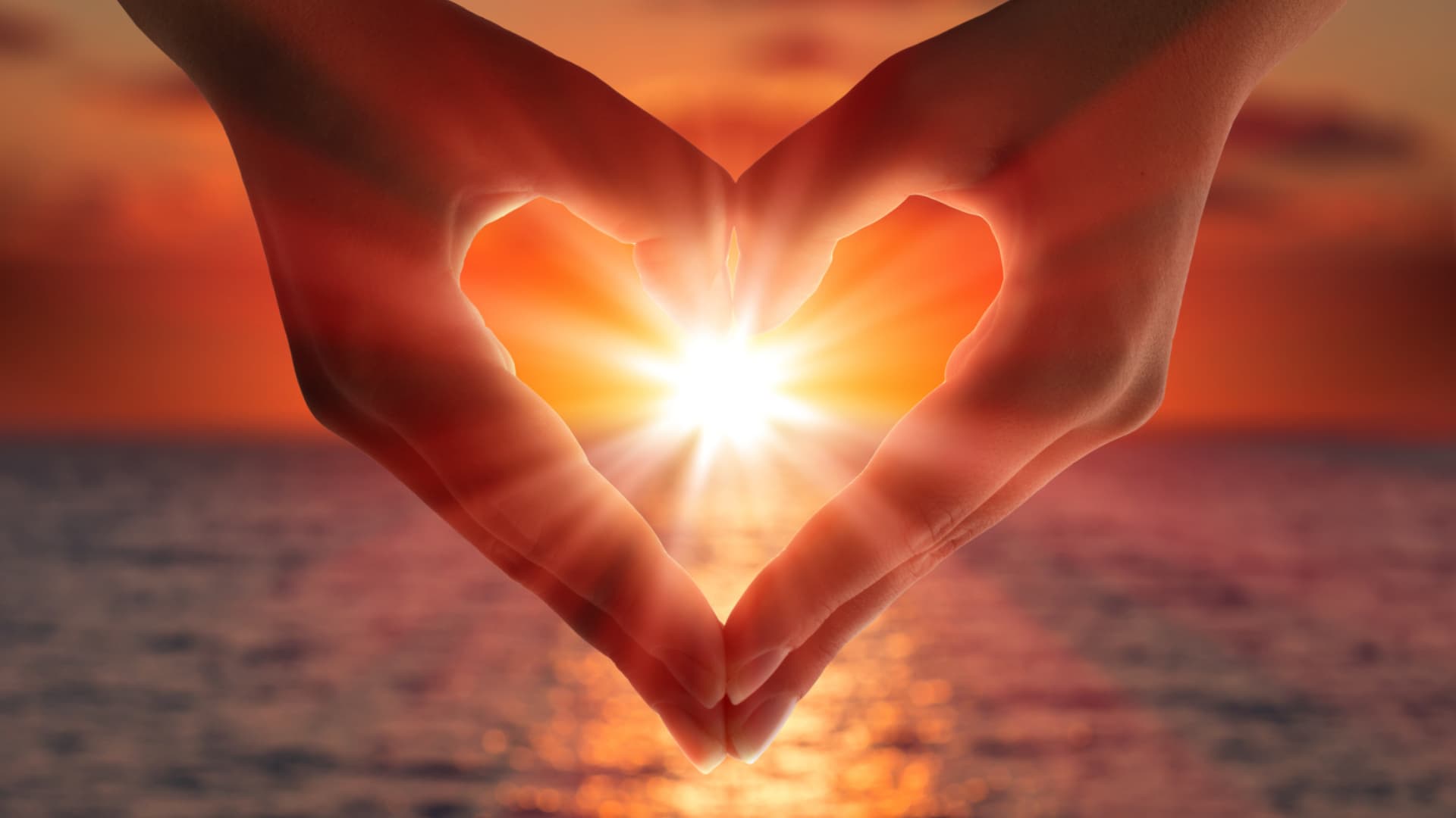 Hands forming a heart shape against a sunset over the ocean.