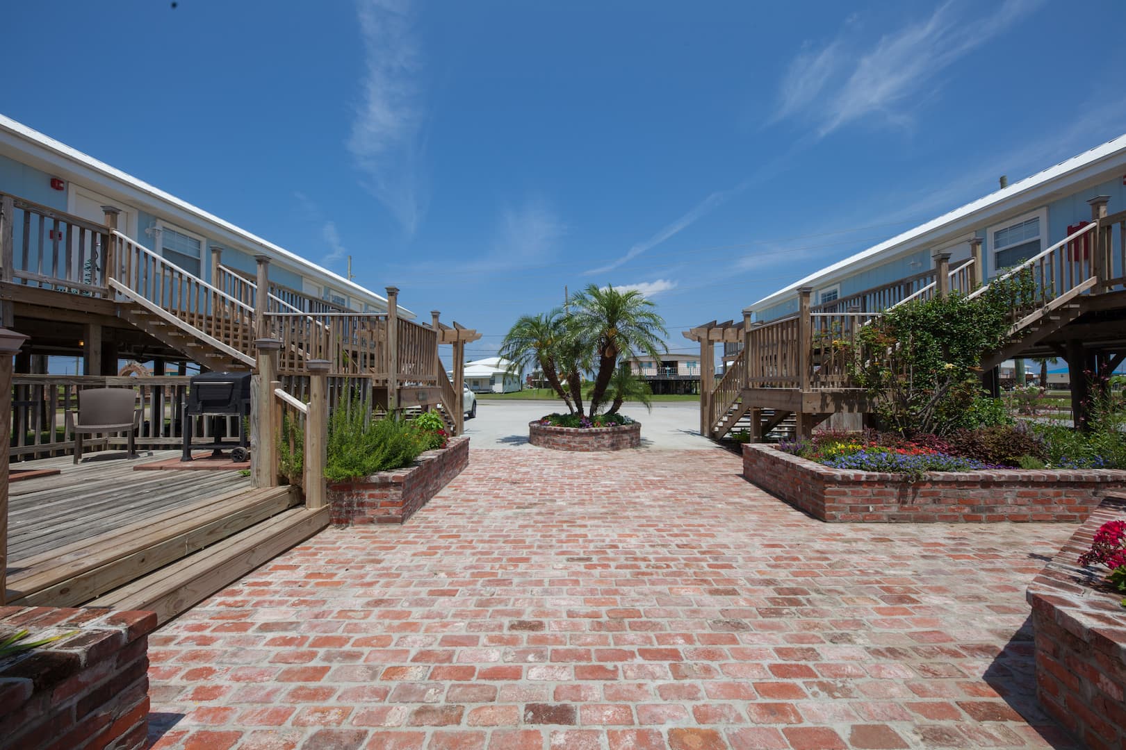 An exterior view of a courtyard between two rows of stilt buildings, featuring a brick path, wooden stairs, and a central palm tree.