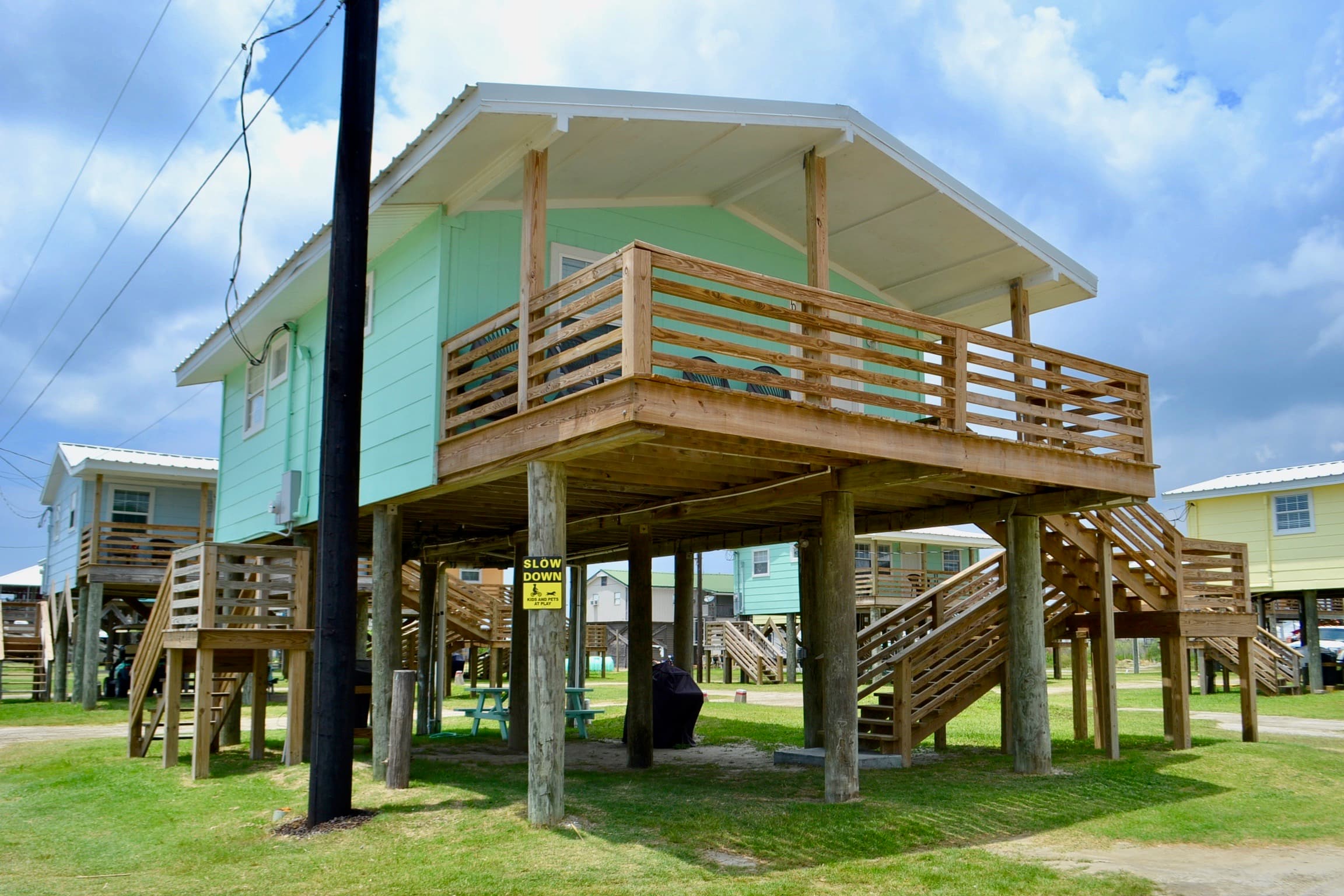 A colorful, elevated mint green stilt house with a wide wooden deck, covered porch, and stairs leading down to a grassy yard.
