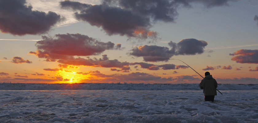 a man standing in the suf of the gulf fishing at sunset